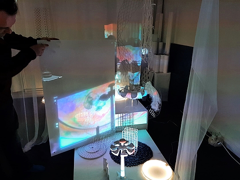 Auf einem Tisch stehen verschiedene Gegenstände und werden mit farbigen Lampen angeleuchtet. Eine Person staht dahinter und hält eine weiße Platte dahinter, sodass die Lichter und die Schattenwürfe darauf zu sehen sind.