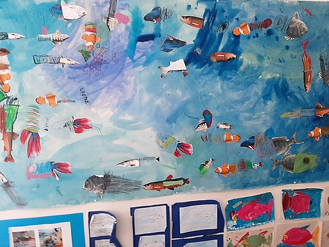 An einer Wand hängt ein großes Stück Papier das mit Wasserfarben und aufgeklebten Motiven von Fischen und anderen Meerestieren eine Unterwasserwelt darstellt